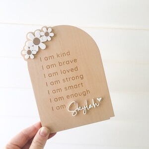Puede incluir: Un letrero de madera con fondo marr&oacute;n claro y texto blanco. El letrero tiene una forma de arco redondeado en la parte superior y presenta un coraz&oacute;n blanco y varias margaritas blancas. El texto dice: "Soy amable, soy valiente, soy amado, soy fuerte, soy inteligente, soy suficiente, soy Skylah".