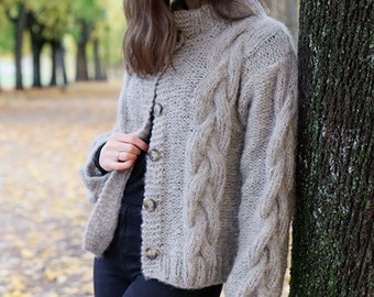 May include: A gray cable knit cardigan with a button closure. The cardigan is worn by a person standing in front of a tree. The text "DROPS DESIGN" and "www.garnstudio.com" is visible at the bottom of the image.