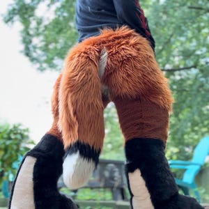 May include: A plush costume featuring a long, thick, orange and brown faux fur tail with a white tip. The lower leg sections are covered in black faux fur with white panels on the inner sides, creating a distinct animal-like appearance.