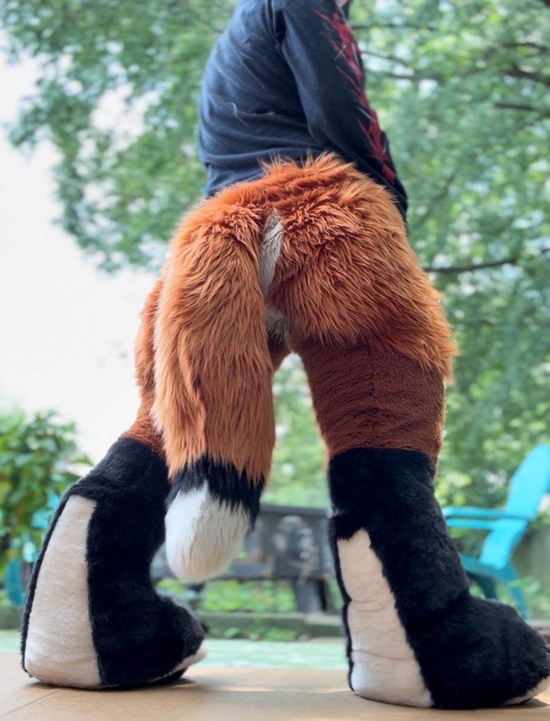 May include: A plush costume featuring a long, thick, orange and brown faux fur tail with a white tip. The lower leg sections are covered in black faux fur with white panels on the inner sides, creating a distinct animal-like appearance.