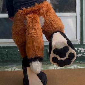 May include: A plush animal costume lower body piece featuring long, shaggy orange faux fur with a white patch on the hip. The costume includes a large, thick orange tail with a white tip and a black base, plus a large, padded black and white paw.