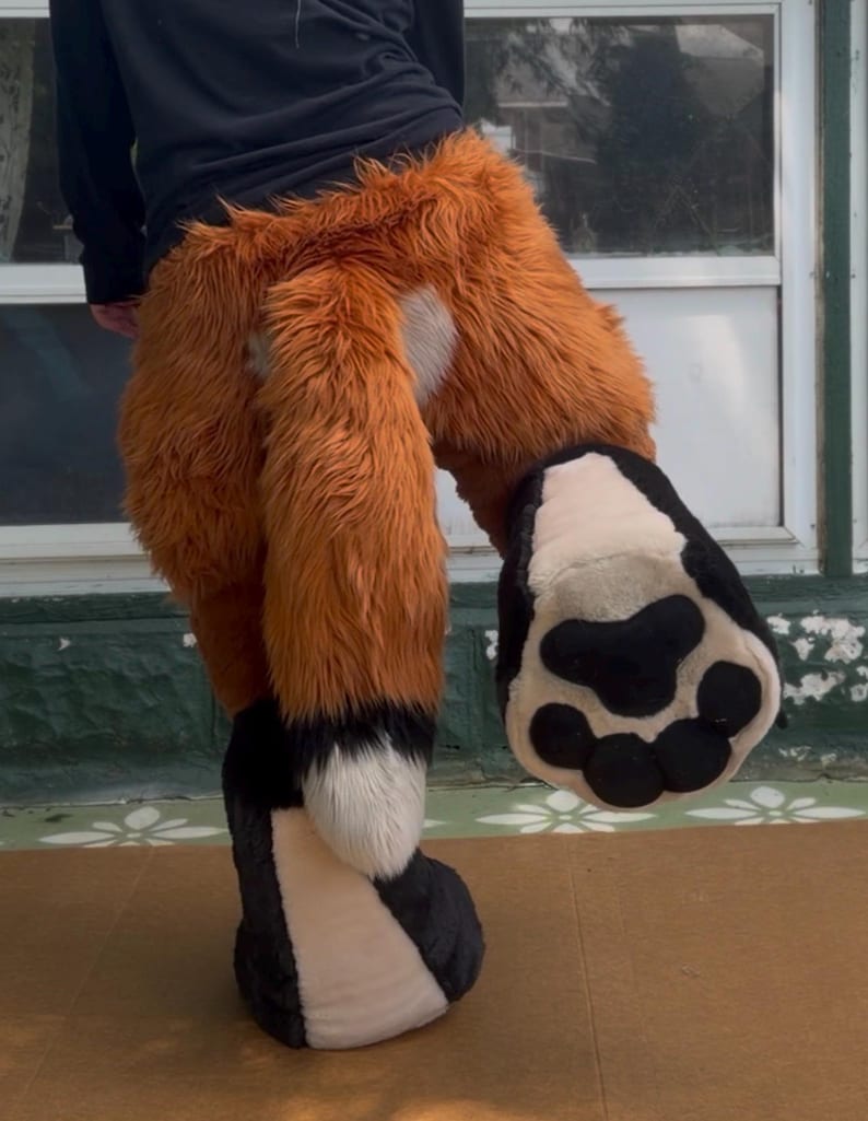May include: A plush animal costume lower body piece featuring long, shaggy orange faux fur with a white patch on the hip. The costume includes a large, thick orange tail with a white tip and a black base, plus a large, padded black and white paw.