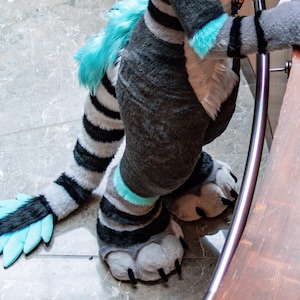 May include: A full-body plush animal costume featuring dark grey fur with white and bright turquoise accents. The costume includes a long, striped tail with a turquoise tuft at the end, large padded paws with black claws, and striped leg sections.
