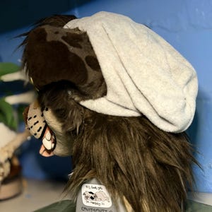 以下が含まれることがあります： 茶色とダークブラウンのフェイクファーで作られた動物のマスクで、上部にはライトグレーのビーニーキャップがかぶせられています。マスクには白い歯が見える鼻先があり、土台には「PLS dont yell im mentally ill CAVITYMUTT」と書かれた小さなステッカーが貼られています。