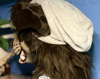 May include: A brown and dark brown faux fur animal mask with a light grey beanie cap covering the top. The mask features a snout with visible white teeth and a small sticker on the base that reads PLS dont yell im mentally ill CAVITYMUTT.