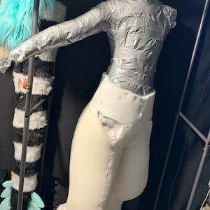 May include: A partial mannequin torso covered in silver duct tape attached to a white foam lower body structure. The upper section features long sleeves and a high neck, while the lower section consists of thick, padded white foam legs.