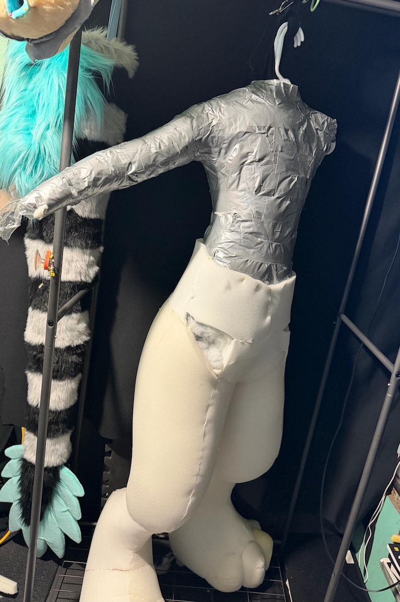 May include: A partial mannequin torso covered in silver duct tape attached to a white foam lower body structure. The upper section features long sleeves and a high neck, while the lower section consists of thick, padded white foam legs.