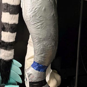 May include: A leg-shaped form covered in overlapping strips of silver duct tape. A blue tape band encircles the ankle area, with black tape stripes above the foot. The text "OUT ANKLE" is written in black ink on the silver tape near the ankle.