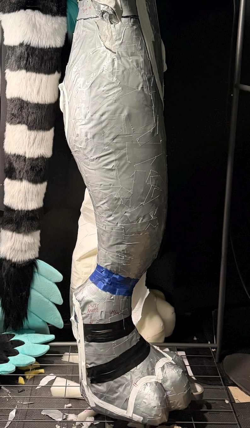 May include: A leg-shaped form covered in overlapping strips of silver duct tape. A blue tape band encircles the ankle area, with black tape stripes above the foot. The text "OUT ANKLE" is written in black ink on the silver tape near the ankle.