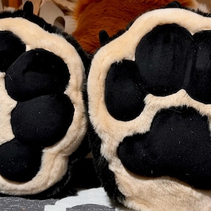 May include: A pair of plush animal paw gloves featuring black velvet-like pads and light beige faux fur trim. The design includes four rounded toe pads and one large central palm pad on each paw, with black fur surrounding the outer edges.