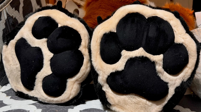 May include: A pair of plush animal paw gloves featuring black velvet-like pads and light beige faux fur trim. The design includes four rounded toe pads and one large central palm pad on each paw, with black fur surrounding the outer edges.
