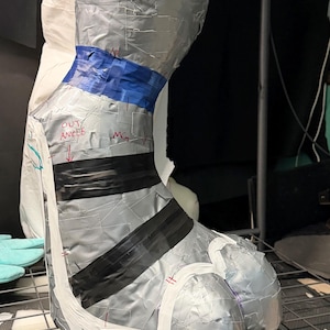 May include: A sculpted leg and foot form covered in silver duct tape, featuring a blue tape band at the ankle, black tape stripes across the foot, and white tape accents. The surface includes handwritten red text reading OUT ANKLE and MG.