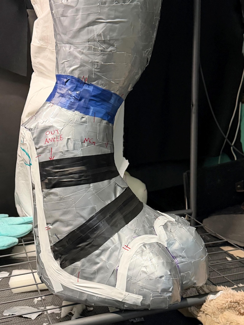 May include: A sculpted leg and foot form covered in silver duct tape, featuring a blue tape band at the ankle, black tape stripes across the foot, and white tape accents. The surface includes handwritten red text reading OUT ANKLE and MG.