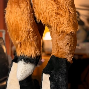 May include: A pair of plush, faux fur leg coverings designed to resemble animal limbs. The upper sections feature long, vibrant orange fur, while the lower portions transition into black fur with white accents on the front and a white patch on the back.