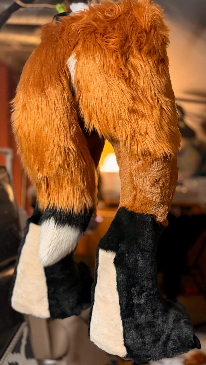 May include: A pair of plush, faux fur leg coverings designed to resemble animal limbs. The upper sections feature long, vibrant orange fur, while the lower portions transition into black fur with white accents on the front and a white patch on the back.