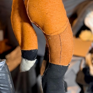 May include: A plush toy fox leg and foot construction made of orange fabric with visible seams. The lower leg is black fabric, ending in a white-tipped paw. The foot is black fabric with stitched details and small yellow accents at the base.