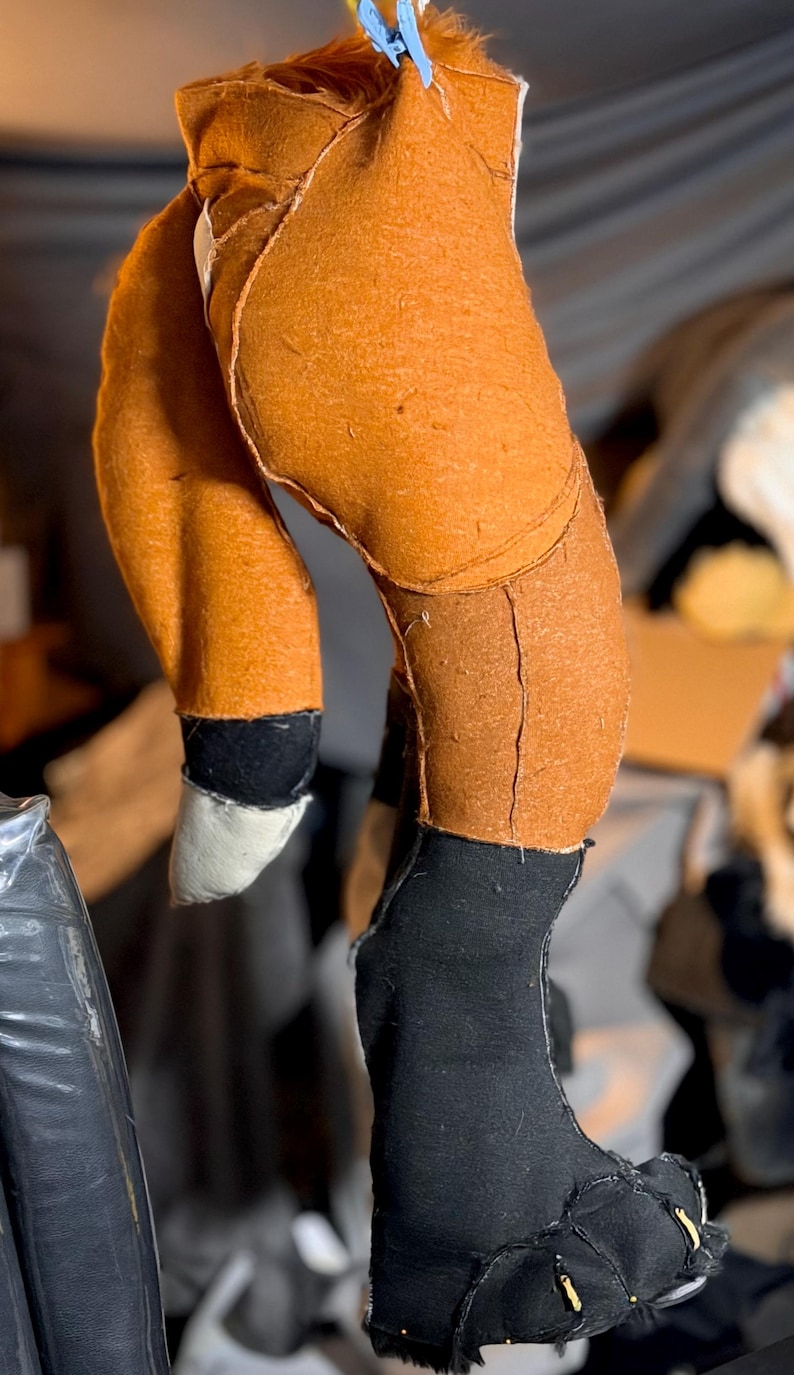 May include: A plush toy fox leg and foot construction made of orange fabric with visible seams. The lower leg is black fabric, ending in a white-tipped paw. The foot is black fabric with stitched details and small yellow accents at the base.