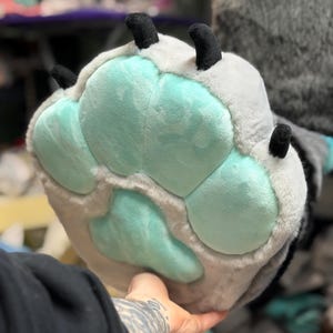 May include: A plush animal paw glove featuring soft light grey faux fur and large, rounded mint green paw pads. The glove includes four distinct black fabric claws extending from the top edge, designed with a thick, padded texture.