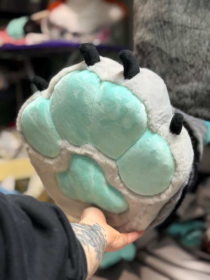May include: A plush animal paw glove featuring soft light grey faux fur and large, rounded mint green paw pads. The glove includes four distinct black fabric claws extending from the top edge, designed with a thick, padded texture.