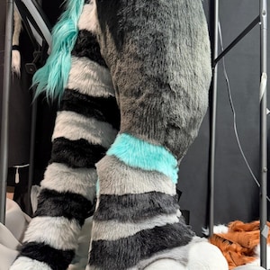 May include: A plush, faux fur animal costume leg and foot piece featuring a grey, black, and white striped pattern with vibrant teal accents. The foot section includes large, padded white toes with black claws and teal paw pads.
