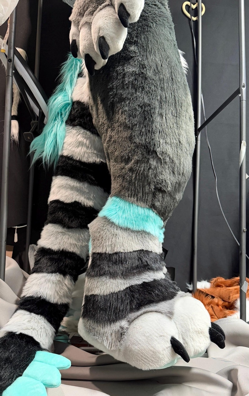 May include: A plush, faux fur animal costume leg and foot piece featuring a grey, black, and white striped pattern with vibrant teal accents. The foot section includes large, padded white toes with black claws and teal paw pads.