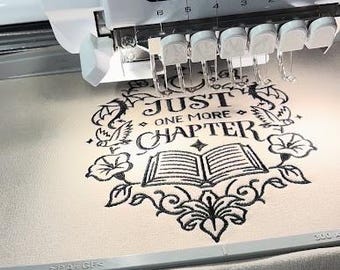 May include: A white Brother Entrepreneur 6-Plus PR670E embroidery machine stitching a black design onto light fabric. The design features an open book, floral elements, and the text 'JUST ONE MORE CHAPTER' enclosed within a decorative circular frame.
