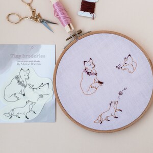Op de afbeelding: Een witte kaart met de tekst "Tiny broderies" en "Les tout petits motifs à broder" en "By Marion Romain" erop gedrukt. De kaart toont een zwart-witte illustratie van drie vossen. Een houten borduurring met een lichtroze stof erin. De stof is geborduurd met drie vossen in bruin.