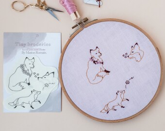 May include: A white card with the text "Tiny broderies" and "Les tout petits motifs à broder" and "By Marion Romain" printed on it. The card shows a black and white illustration of three foxes. A wooden embroidery hoop with a light pink fabric inside. The fabric has brown embroidery of three foxes.