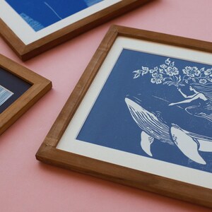 May include: Three framed art prints with wooden frames. One print features a whale with a woman and flowers on a blue background. The other two prints have abstract blue and white designs. The frames are a warm brown color.