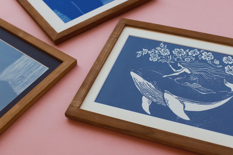 May include: Three framed art prints with wooden frames. One print features a whale with a woman and flowers on a blue background. The other two prints have abstract blue and white designs. The frames are a warm brown color.