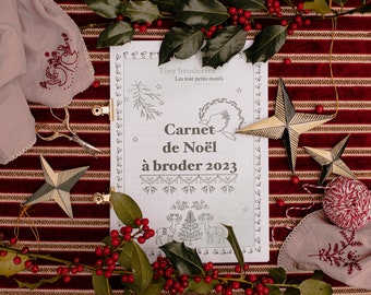 May include: A white booklet with the title "Carnet de Noël à broder 2023" and the text "Tiny broderies Les tout petits motifs" on the cover. The booklet is surrounded by red and white striped fabric, holly sprigs with red berries, and gold star ornaments.
