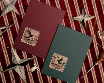 May include: Two cards with a dark red and dark green background. Each card has a square cutout with a brown background and a black silhouette of a bird flying over a body of water with trees in the background.