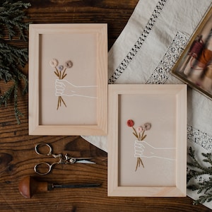 Könnte beinhalten: Zwei gerahmte Stickbilder mit einer Hand, die einen Blumenstrauß hält. Die Blumen sind aus Garn gefertigt und in den Farben Rosa, Rot und Lila gehalten. Der Hintergrund der Stickerei ist hellbeige.