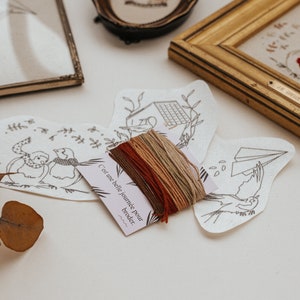 Peut inclure: Trois motifs de broderie représentant un oiseau avec un avion en papier, deux oiseaux dans un cercle de feuilles et un oiseau dans un nichoir. Les motifs sont imprimés sur un tissu blanc et sont accompagnés d'une pelote de fil à broder dans des tons de brun, orange et rouge. Le fil est étiqueté "C'est une belle journée pour broder."
