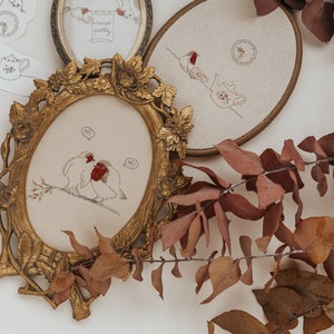 Peut inclure: Deux cerceaux brodés avec un motif floral. Un cerceau représente une paire d'oiseaux avec des accents rouges et le texte "Salut !" dans une bulle de dialogue. L'autre cerceau représente un oiseau avec un accent rouge et une théière.