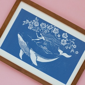 May include: A framed art print featuring a white illustration on a blue background. The artwork depicts a woman riding a whale, holding flowers. The print is in a wooden frame with a white mat, set against a pink background.