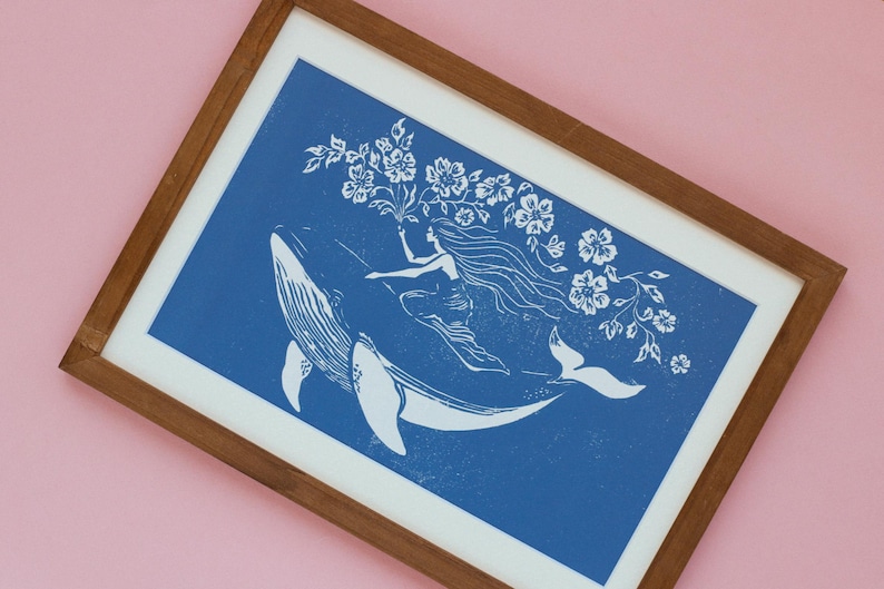 May include: A framed art print featuring a white illustration on a blue background. The artwork depicts a woman riding a whale, holding flowers. The print is in a wooden frame with a white mat, set against a pink background.