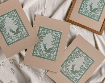 May include: Four brown paper prints with a green leafy design and a bird silhouette in the center. The prints are on a white fabric background.