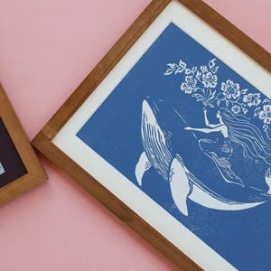 May include: Two framed art prints. One features a blue and white illustration of a woman riding a whale, holding flowers. The other print has blue and white abstract lines. Both are in wooden frames, set against a pink background.