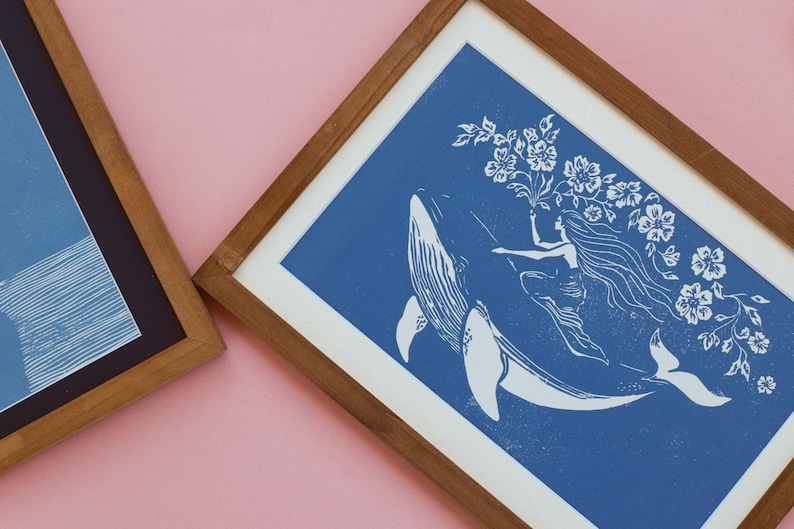 May include: Two framed art prints. One features a blue and white illustration of a woman riding a whale, holding flowers. The other print has blue and white abstract lines. Both are in wooden frames, set against a pink background.