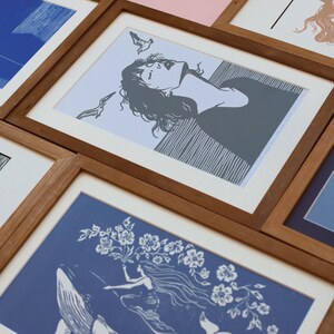 May include: A collection of framed art prints with wooden frames. The prints feature illustrations in shades of blue, gray, and tan. The artwork depicts figures, flowers, and abstract designs. The frames are arranged at various angles, creating a visually appealing display.