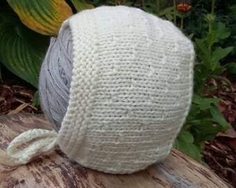 May include: A white knitted baby bonnet with a tie. The bonnet is sitting on a piece of wood.