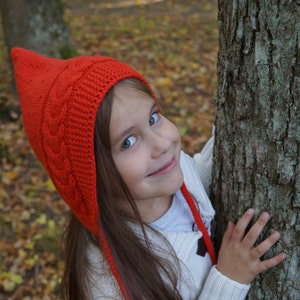 Peut inclure: Une jeune fille portant un bonnet rouge tricoté avec une longue pointe et un pull blanc, souriant et regardant la caméra. Elle est debout à côté d'un tronc d'arbre.