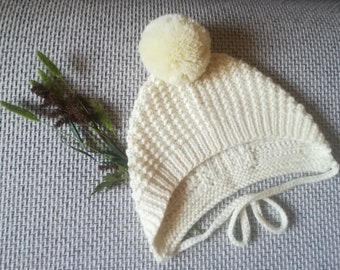 May include: A white knitted baby hat with a white pom pom on top. The hat has a tie at the bottom.