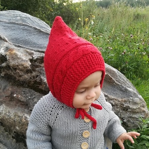 May include: A baby wearing a red knitted bonnet with a pointed top and a grey knitted cardigan with wooden buttons.