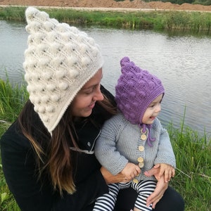 May include: A woman and a child wearing matching knitted hats with a textured bobble design. The woman's hat is white and the child's hat is purple. The child is wearing a grey knitted cardigan with wooden buttons.
