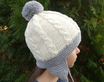 Puede incluir: Un gorro de punto blanco y gris con un pompón en la parte superior. El gorro tiene un patrón de punto de cable y orejeras.