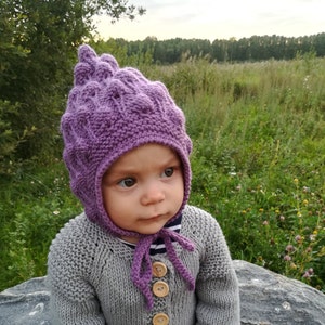 Op de afbeelding: Een baby draagt ​​een paarse gebreide muts met een puntige top en een grijze gebreide trui met houten knopen.
