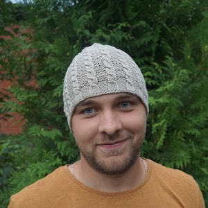 May include: A person wearing a light beige knitted hat with a cable knit pattern. The hat is pulled down over their forehead and covers their ears. The person is smiling and looking at the camera.