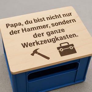 May include: A blue storage crate with a light wood lid. The lid has the text "Papa, du bist nicht nur der Hammer, sondern der ganze Werkzeugkasten" along with a hammer and toolbox graphic.